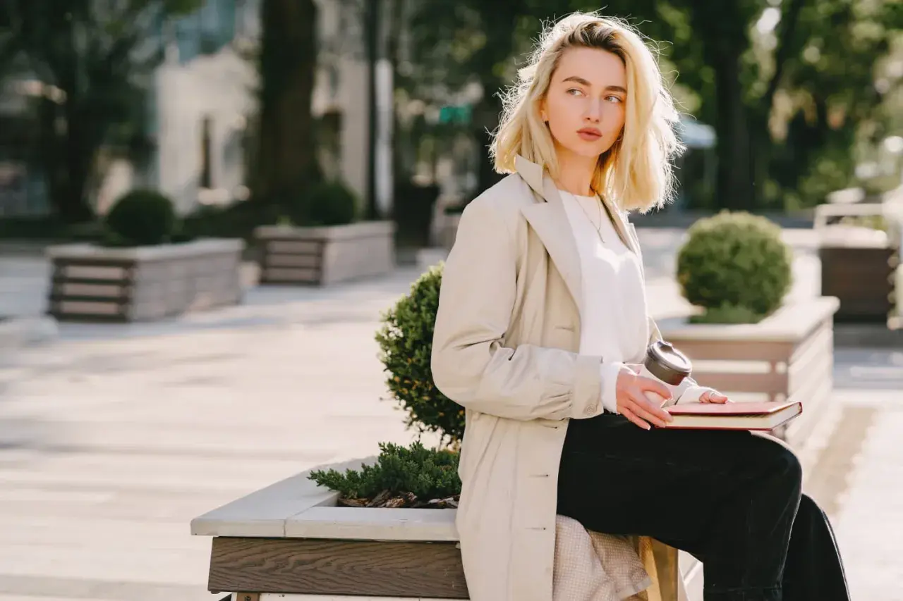 Une femme blonde, assise dehors avec un livre et un café, porte un trench beige et un haut blanc. Parfait pour savoir comment s'habiller quand il fait 15 degrés.