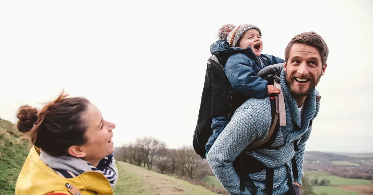 Szczęśliwa rodzina na spacerze. Tata niesie dziecko w nosidełku turystycznym, mama uśmiecha się do nich.