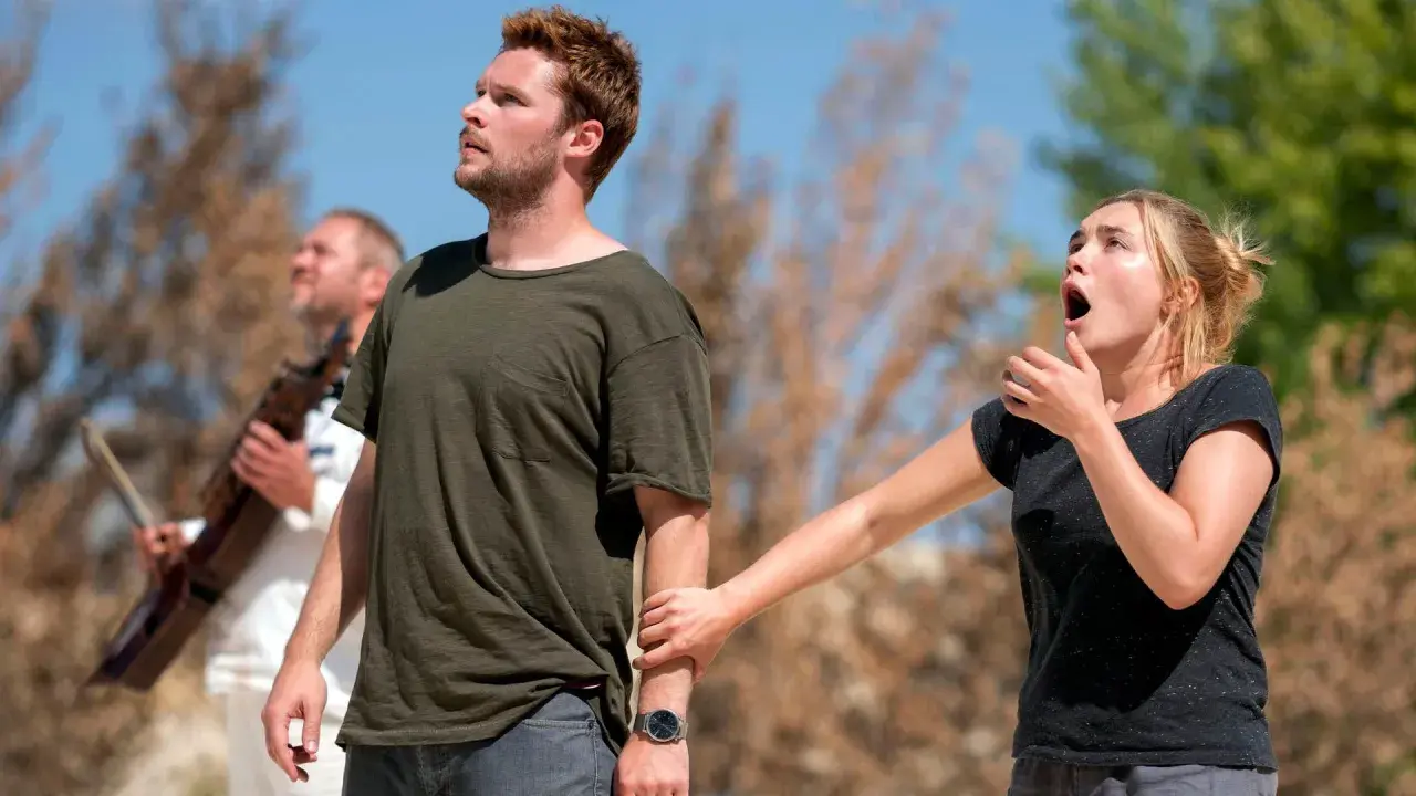 En kvinna griper tag i en mans arm, båda ser uppåt med förvånade uttryck. En man med ett instrument syns i bakgrunden. Scen från Midsommar filmens rollista.