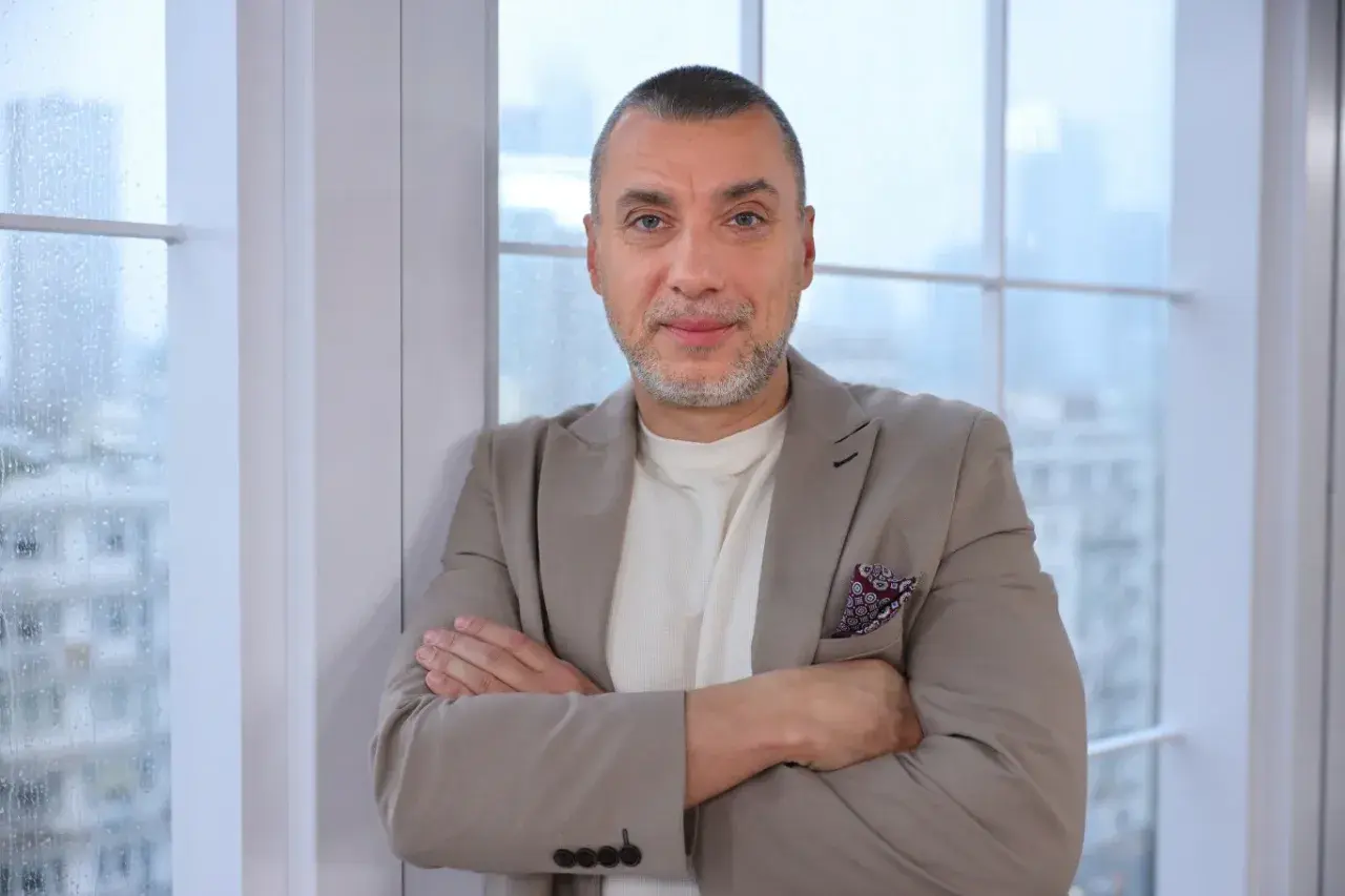Maciej Dębosz prywatnie, w beżowej marynarce i z dumnym spojrzeniem, opiera się o okno.