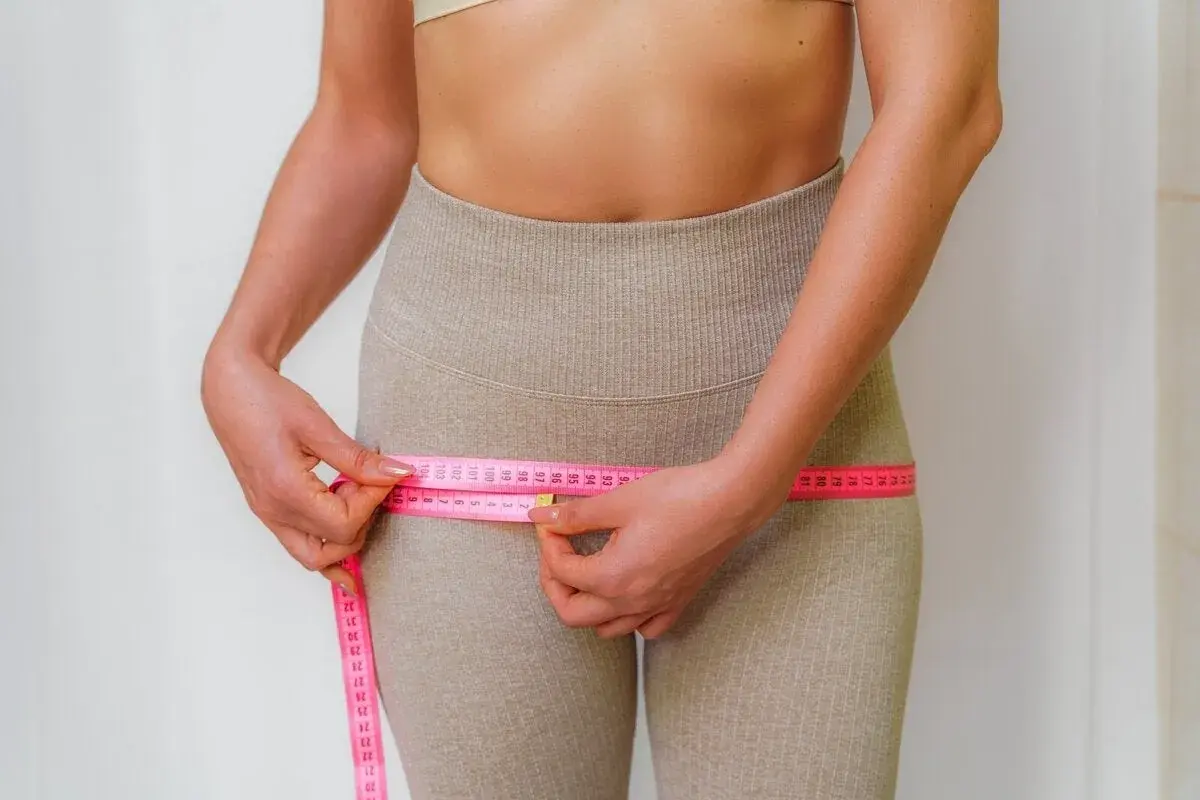 Mujer midiendo su cintura, observando la cinta métrica rosa que rodea sus caderas, enfocándose en los contornos y posibles hip dips.