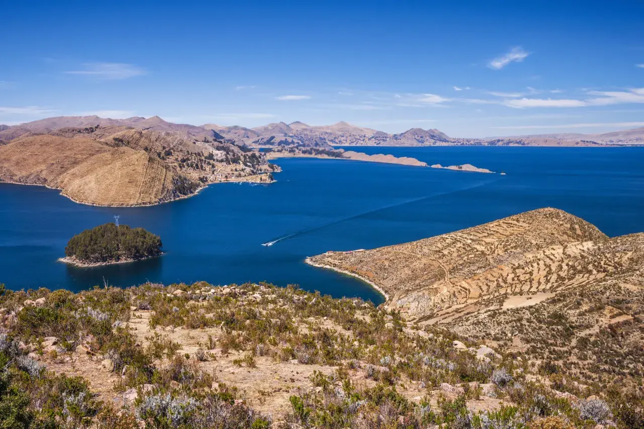 Blick auf den Titicacasee, wo die Anden auf das tiefblaue Wasser treffen. Terrassierte Hänge und eine kleine Insel mit Bäumen zeugen von der Schönheit dieser Region.