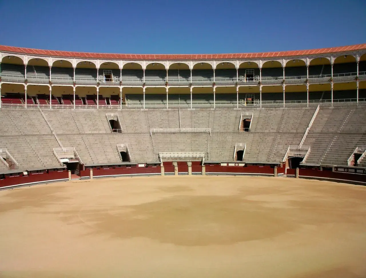 Vista interior de una plaza de toros vacía, con gradas de piedra y balcones arqueados. Las ventas por dentro esperan al público.