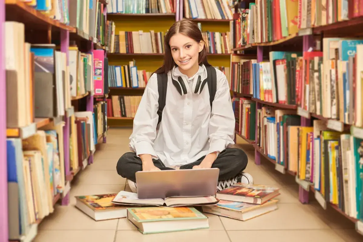 Dziewczyna z laptopem i słuchawkami siedzi na podłodze w bibliotece, otoczona regałami z książkami.