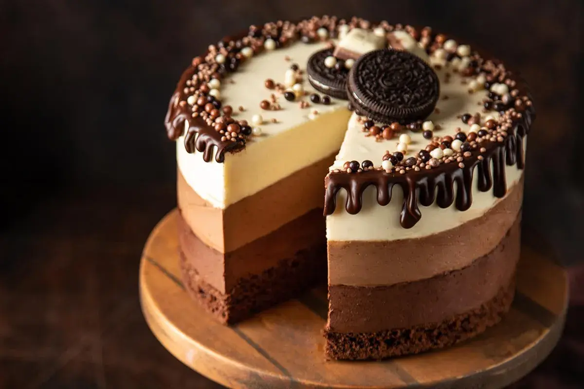 Torta ai tre cioccolati ricetta facile: golosa e scenografica, con strati di mousse al cioccolato bianco, al latte e fondente, decorata con gocce di cioccolato e biscotti.