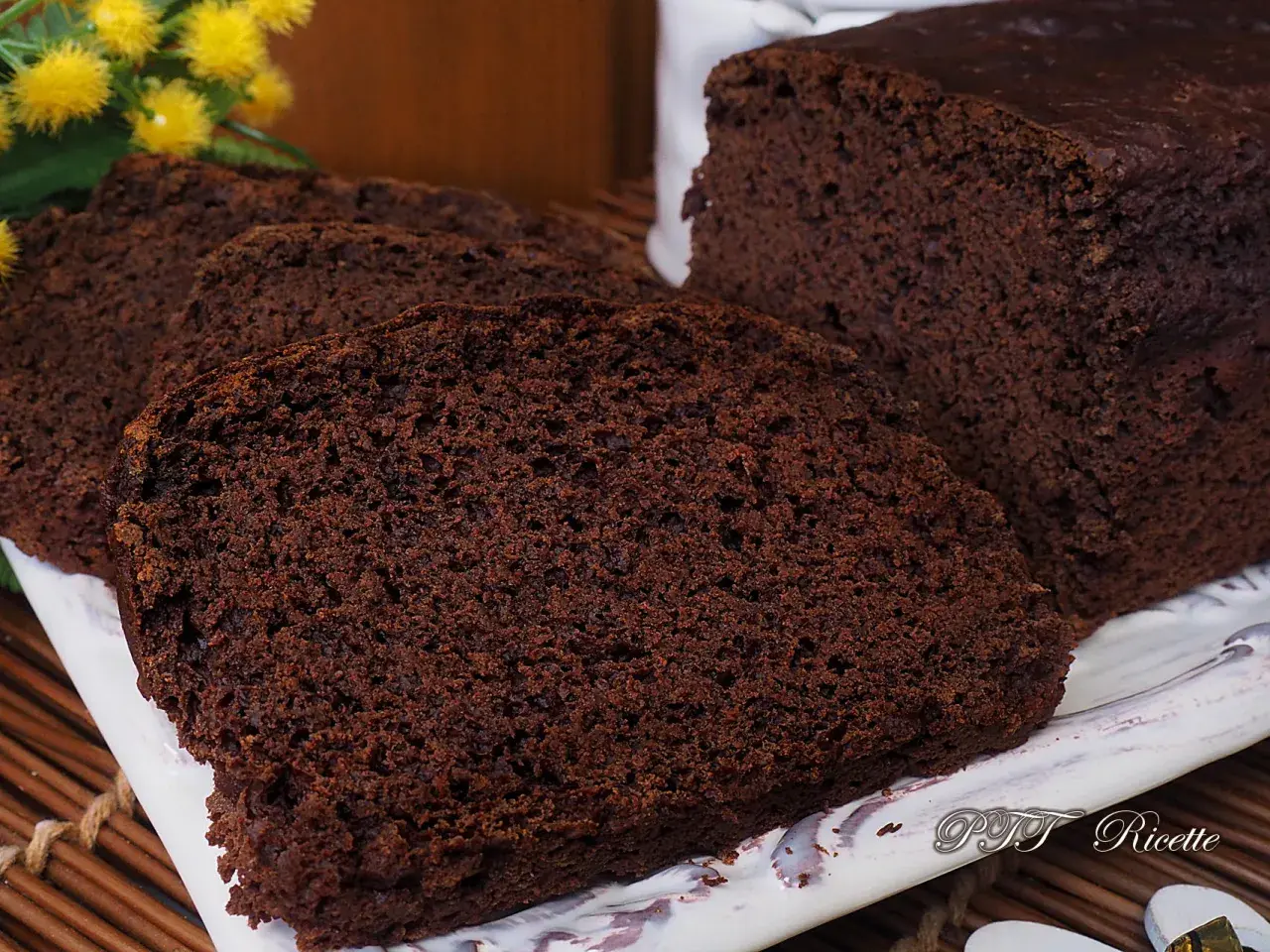 Plumcake al cacao sofficissimo, affettato su un piatto bianco, con fiori gialli sullo sfondo.