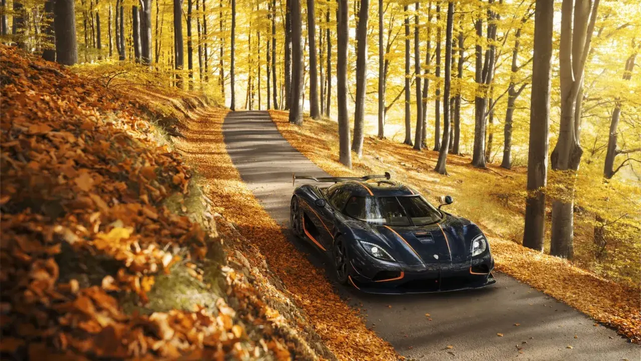 Mroczny Koenigsegg Agera RS z pomarańczowymi akcentami przemierza jesienną drogę wśród złotych liści i drzew.