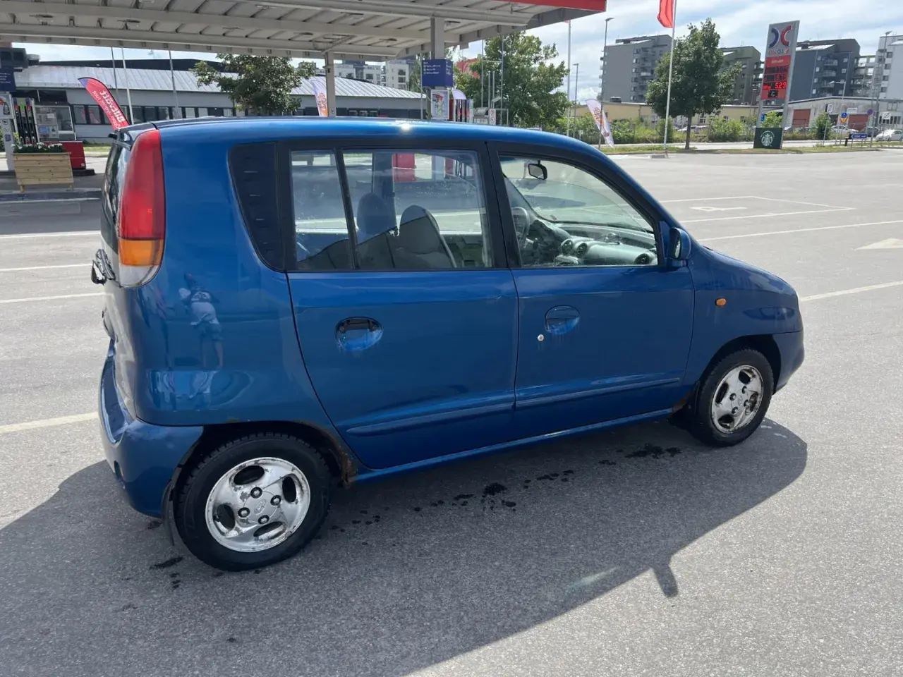 En blå Hyundai Atos står parkerad vid en bensinstation. Bilen har några rostfläckar.
