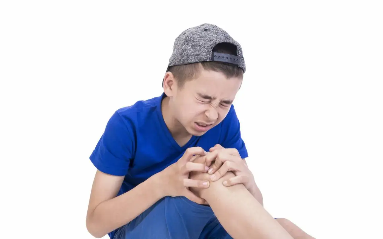 Ragazzo con cappellino grigio stringe il ginocchio con espressione sofferente, forse a causa dei dolori della crescita.