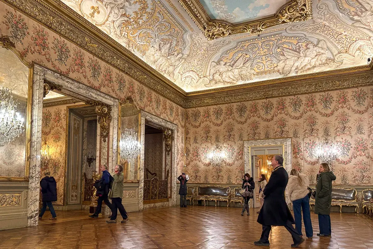 Elegancki przedpokój w pałacu z bogato zdobionym sufitem i tapetą w kwiaty. Zwiedzający podziwiają wnętrze.