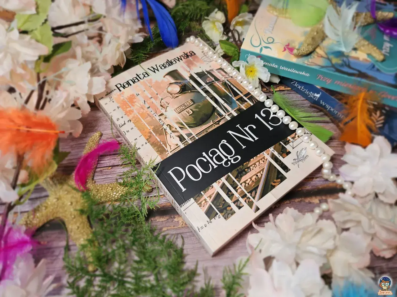 Książka "Pociąg Nr 13" Renaty Wasilewskiej, ozdobiona perłami i piórami, niczym oryginał oprócz błękitnego nieba, otoczona kwiatami.