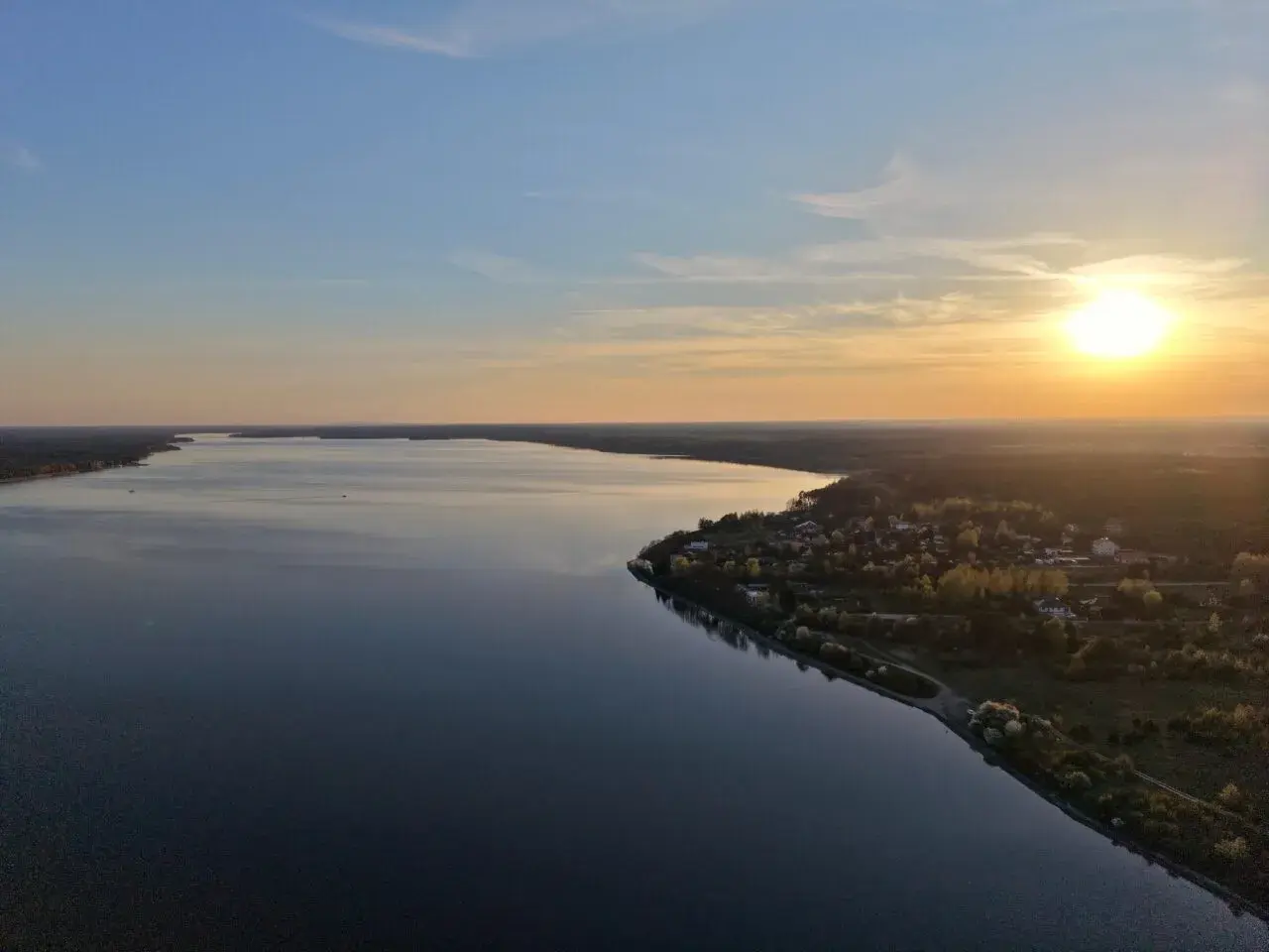 Zachód słońca nad Zalewem Sulejowskim. Spokojna tafla wody odbija niebo, a na brzegu widać domki letniskowe i las.