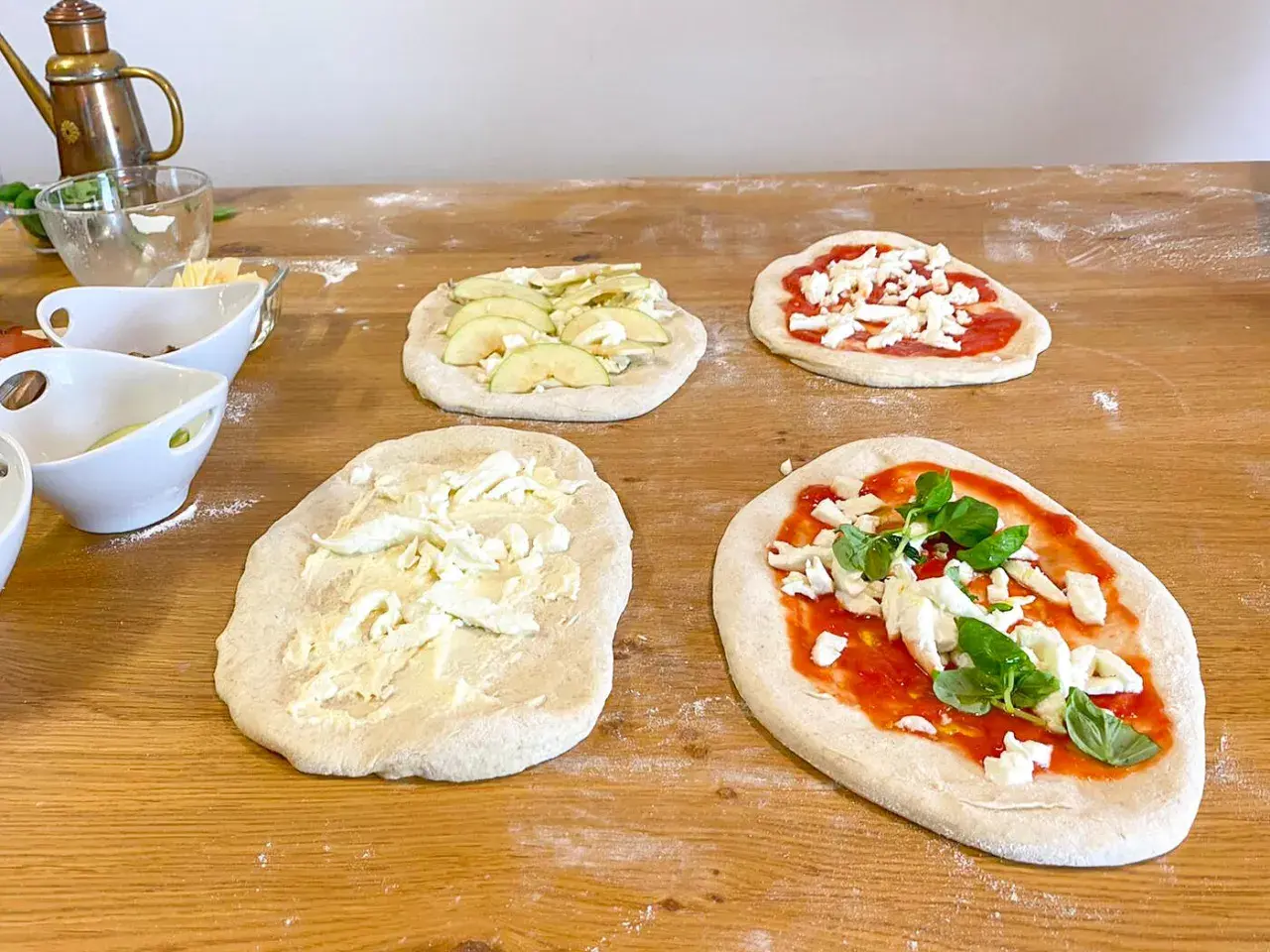 Vier rohe Pinsa-Teige auf einem Holztisch, teils belegt mit Tomatensoße, Mozzarella, Äpfeln und Basilikum.