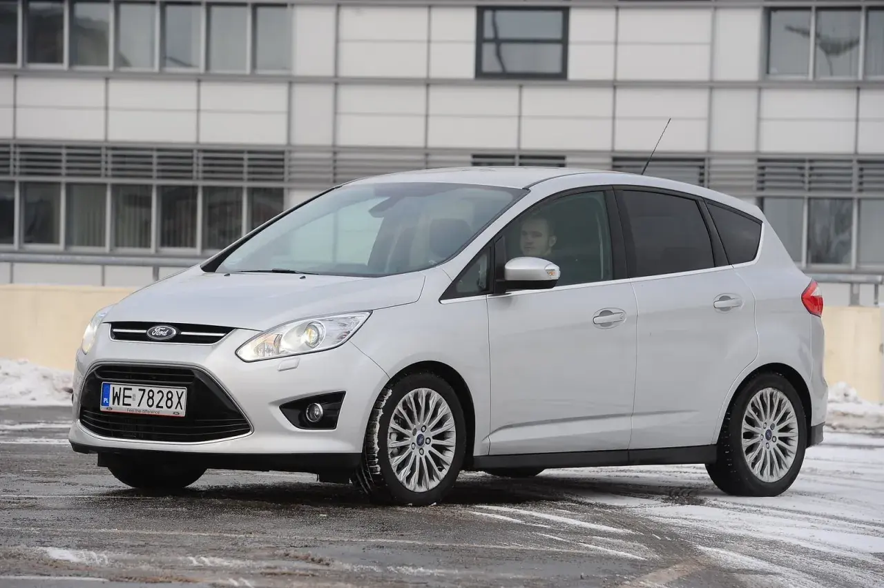 Srebrny Ford C-Max z włączonymi przednimi światłami, gotowy do jazdy w zimowych warunkach.