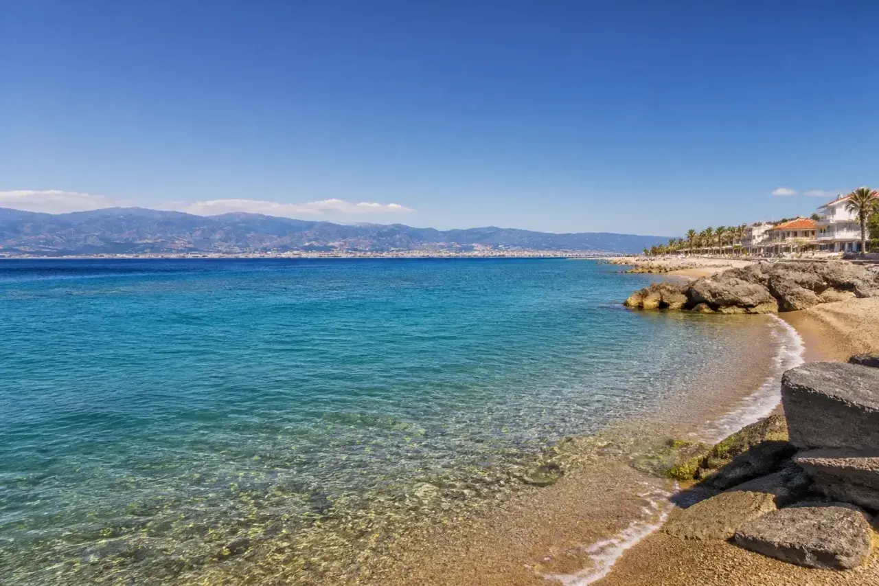 Spiaggia di Reggio Calabria con mare cristallino, palme e case bianche. All'orizzonte, montagne azzurre sotto un cielo limpido.