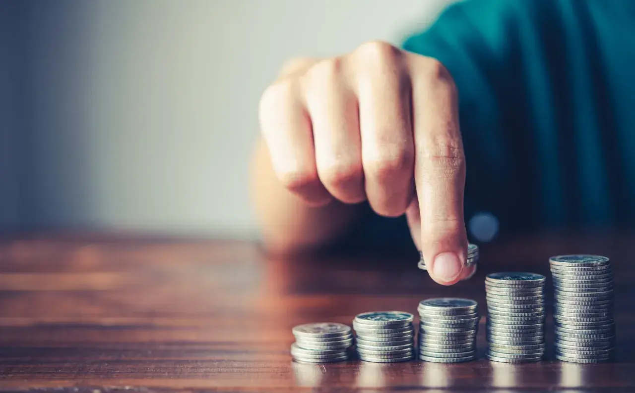 Mano che aggiunge una moneta a una pila, simboleggiando la crescita finanziaria, il risparmio o la gestione del **tutore e indennità di accompagnamento**.