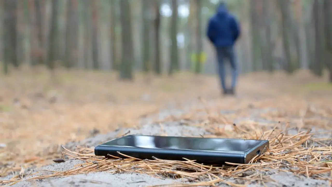 Ein Handy verloren, was tun? Ein schwarzes Smartphone liegt im Wald auf Nadeln. Im Hintergrund geht eine Person weg.