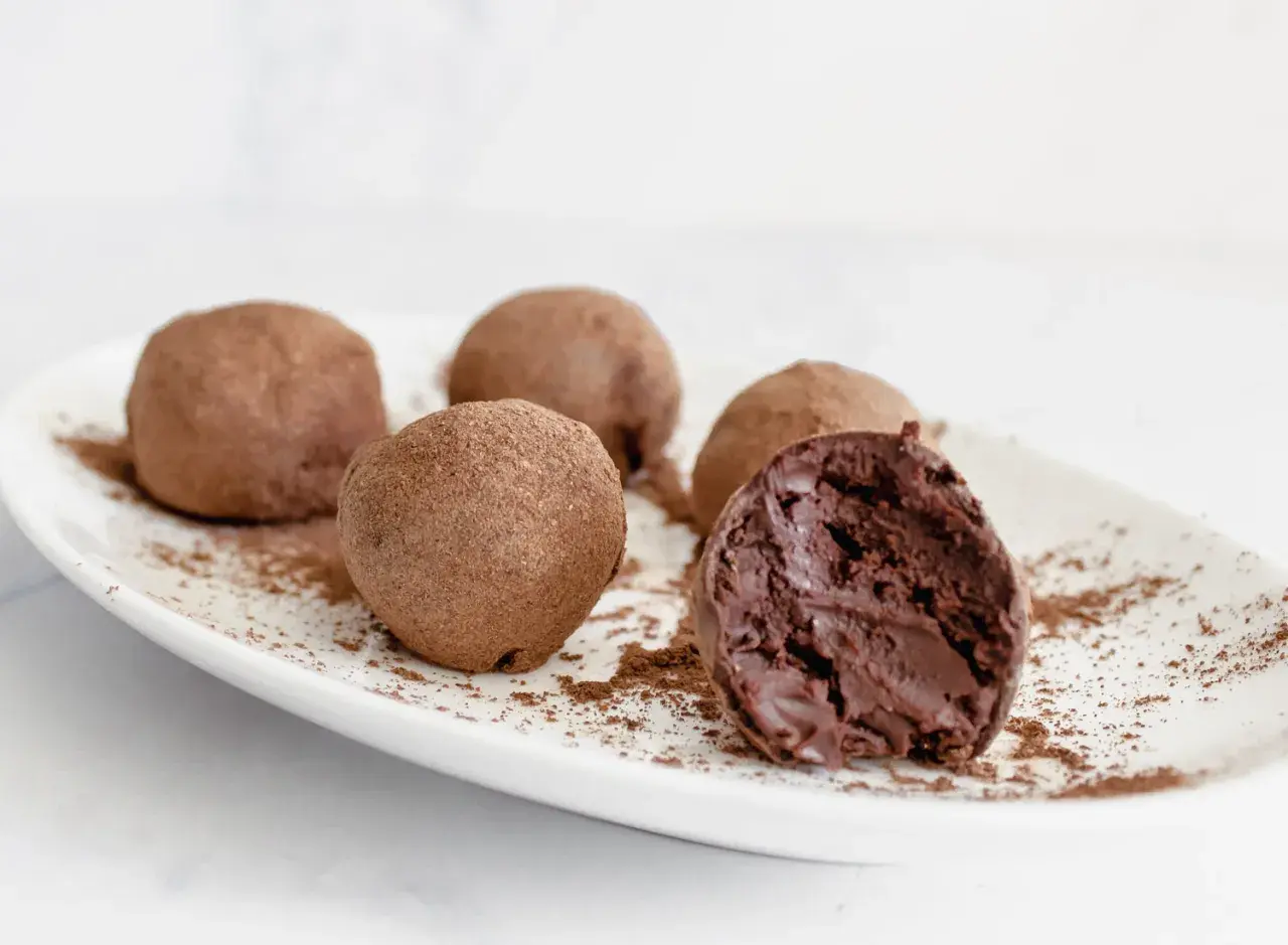 Cinco trufas de chocolate espolvoreadas con cacao en polvo sobre un plato blanco. Una trufa está partida, revelando su interior cremoso.
