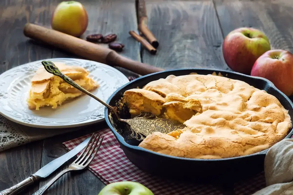 Una deliziosa torta di mele in padella, appena sfornata, con una fetta servita su un piatto. Mele fresche e cannella completano la scena rustica.