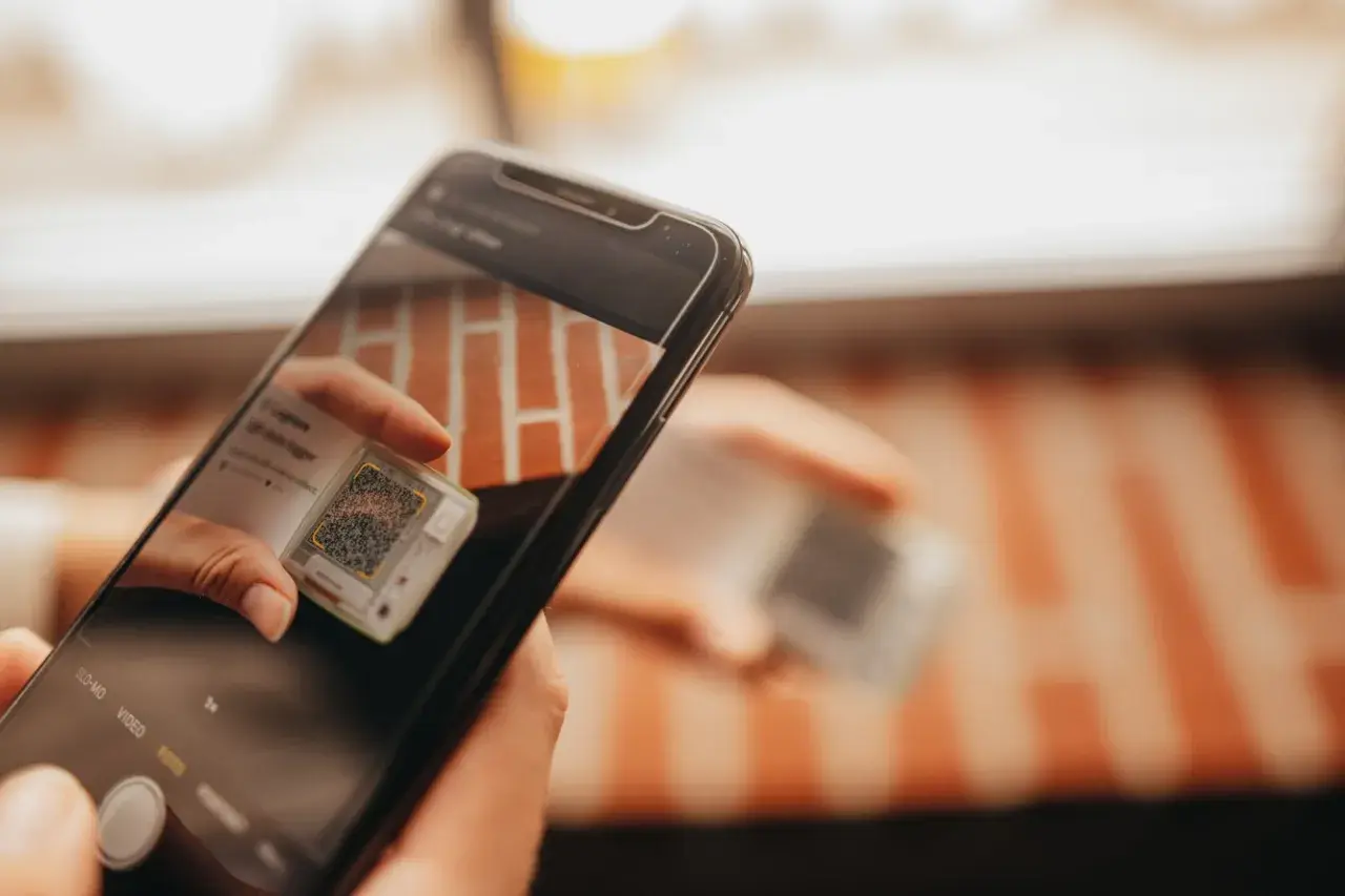 Ręce trzymają smartfon, który skanuje kod QR na małym urządzeniu. To może być część procesu śledzenia przesyłki przez kuriera Hellmann.