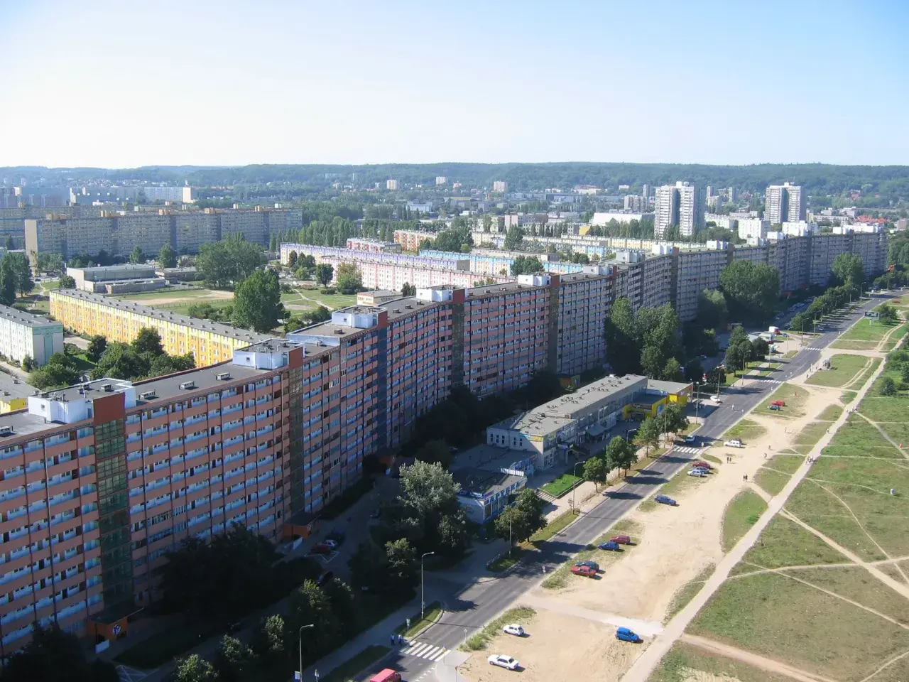 Długi, falisty budynek mieszkalny, znany jako falowiec gdańsk, rozciąga się wzdłuż ulicy, otoczony zielenią i innymi blokami.