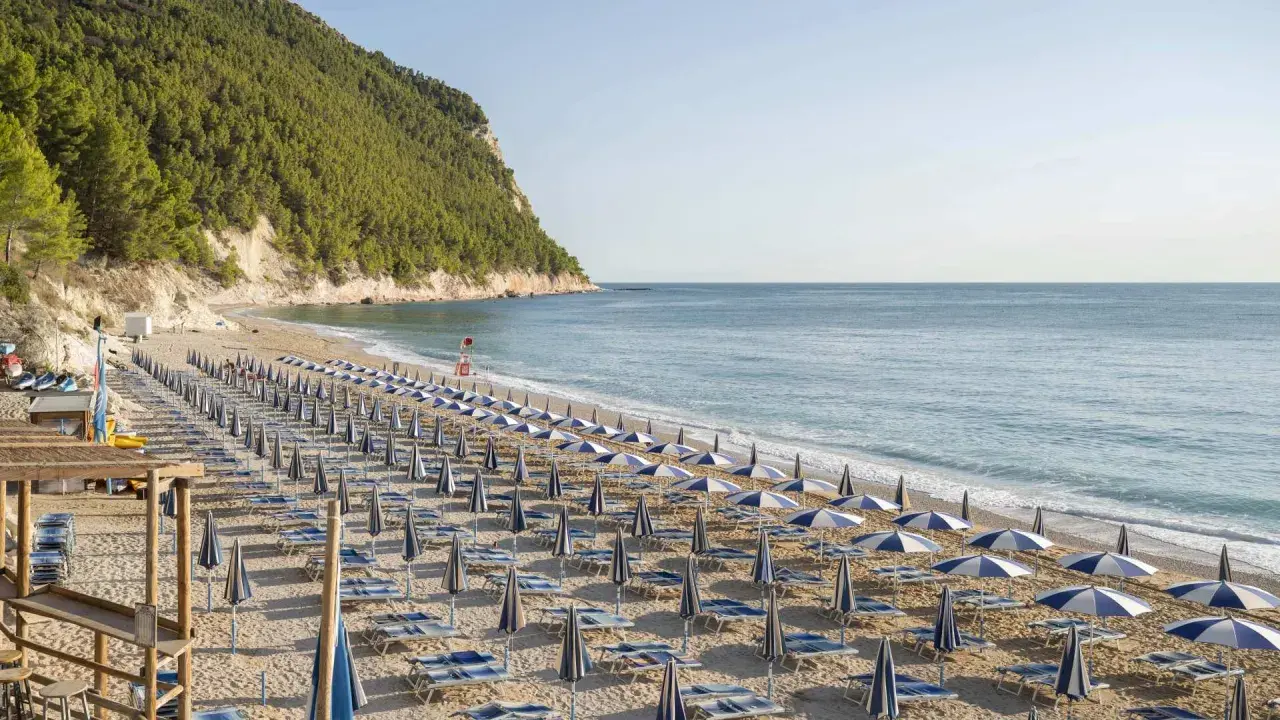 Filettata di ombrelloni e lettini sulla spiaggia di San Michele, con il mare azzurro e la collina verde sullo sfondo.