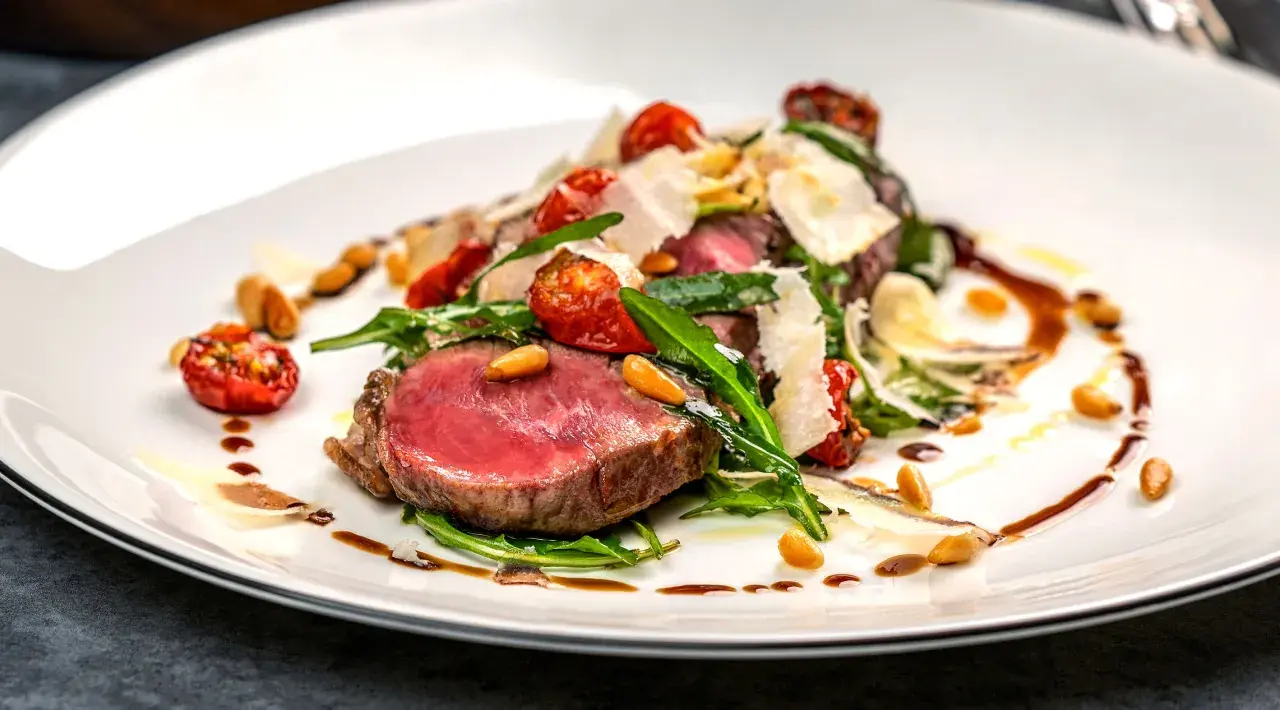 Saftiges Tagliata di Manzo mit Rucola, Kirschtomaten, Pinienkernen und Parmesan auf einem weißen Teller.