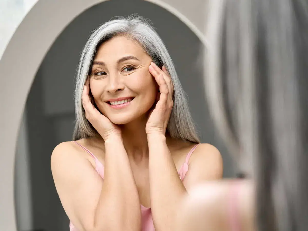 Une femme souriante aux cheveux gris, se touchant le visage, cherche à combler les rides naturellement.