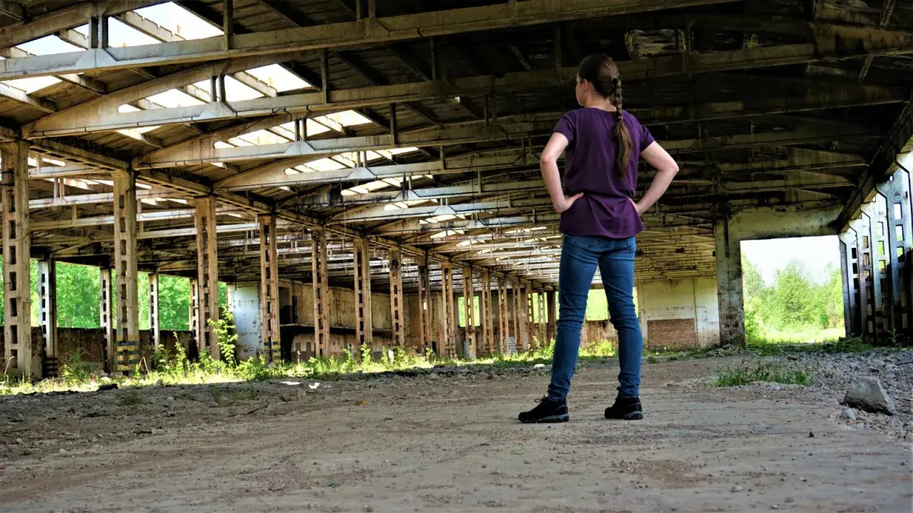 Dziewczynka w jeansach i fioletowej bluzce stoi w opuszczonej hali, odkrywając tajemnice urbex.