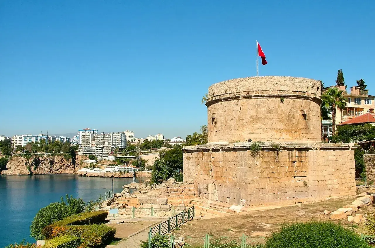 Antalya Altstadt: Ein runder Steinturm mit türkischer Flagge überragt die Bucht. Im Hintergrund moderne Gebäude und Felsen.