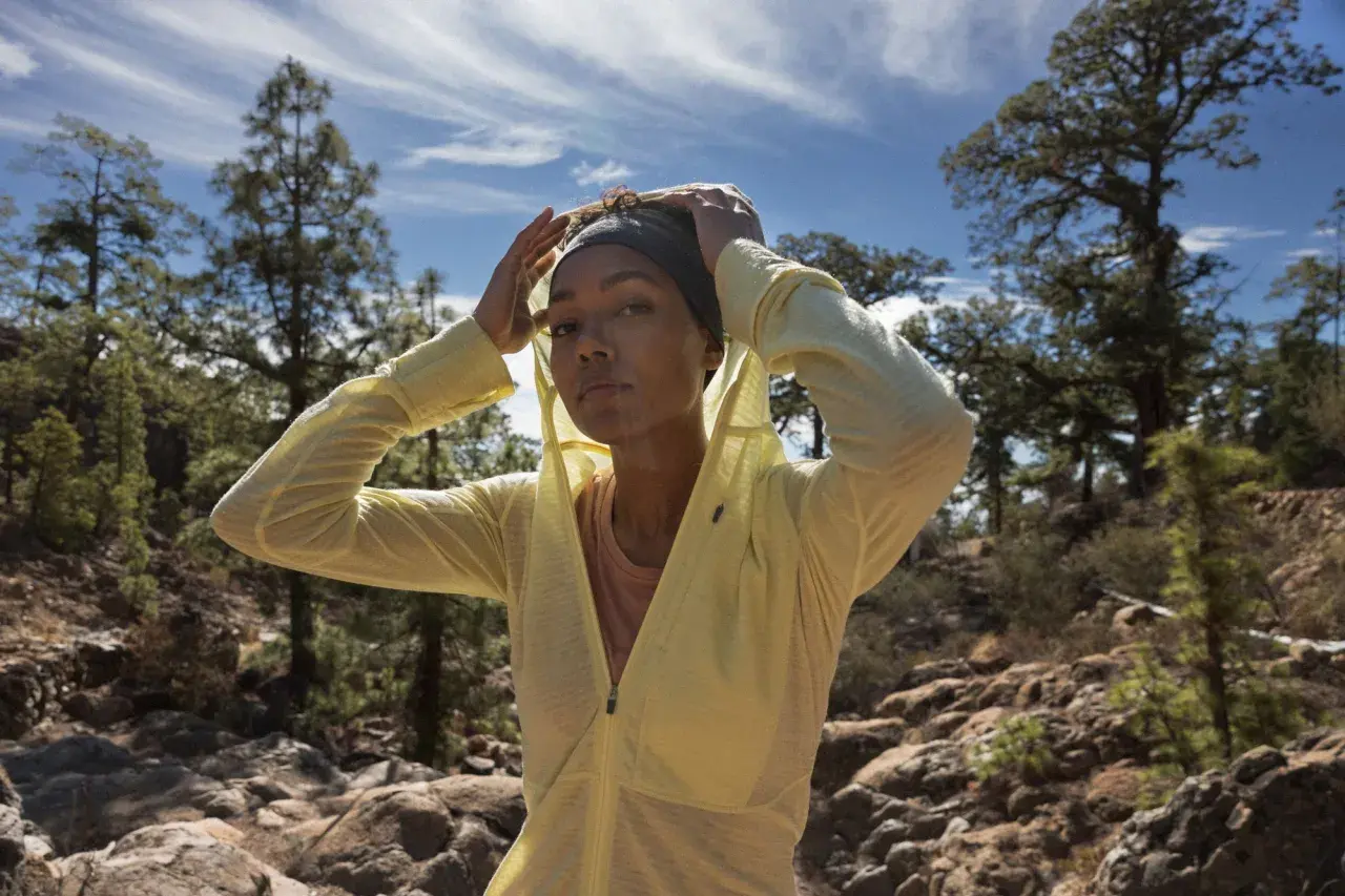 Une femme ajuste son bandeau et sa veste légère, parfaite pour savoir comment s'habiller pour une randonnée en été. Paysage rocheux et arbres en arrière-plan.