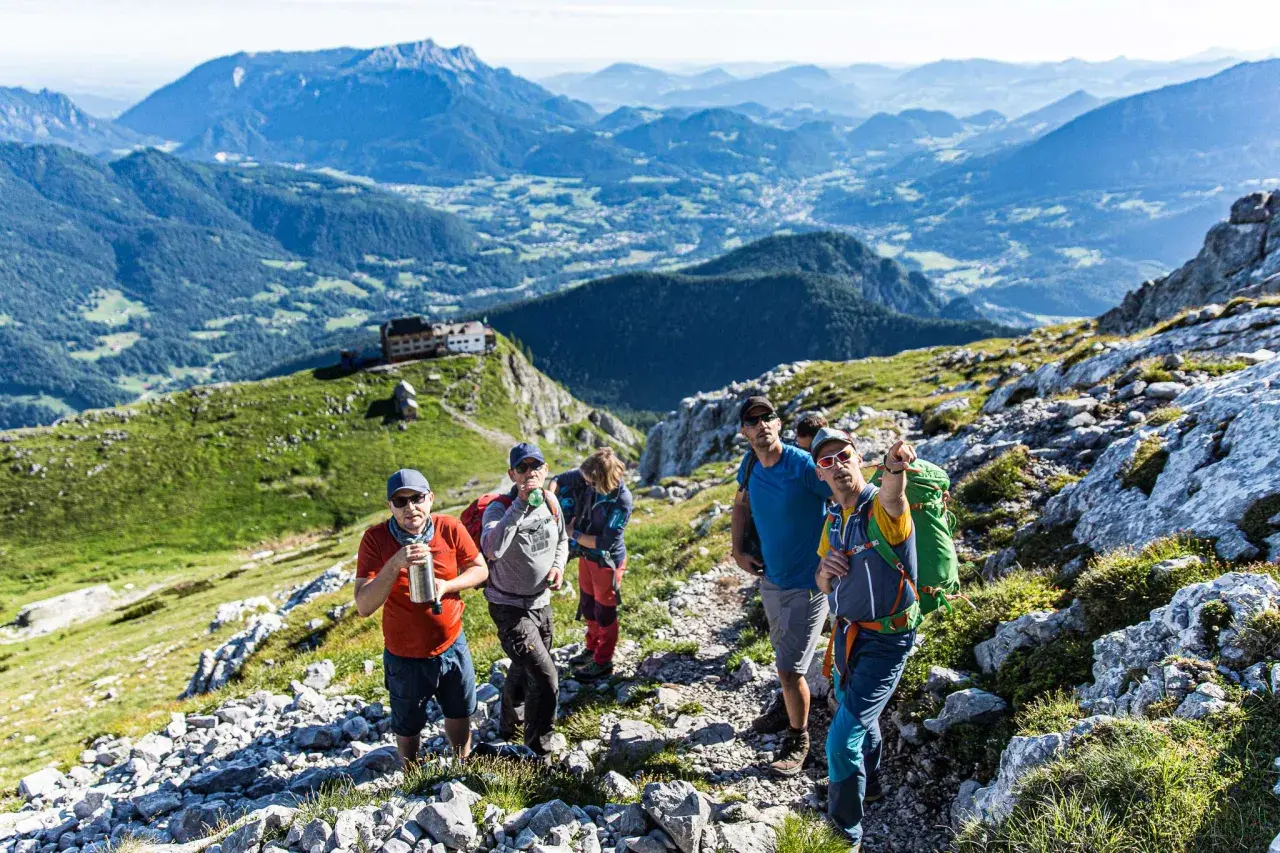 Wandergruppe auf dem Weg zum hocheck Watzmann. Im Hintergrund ein Alpenpanorama mit einem Dorf.