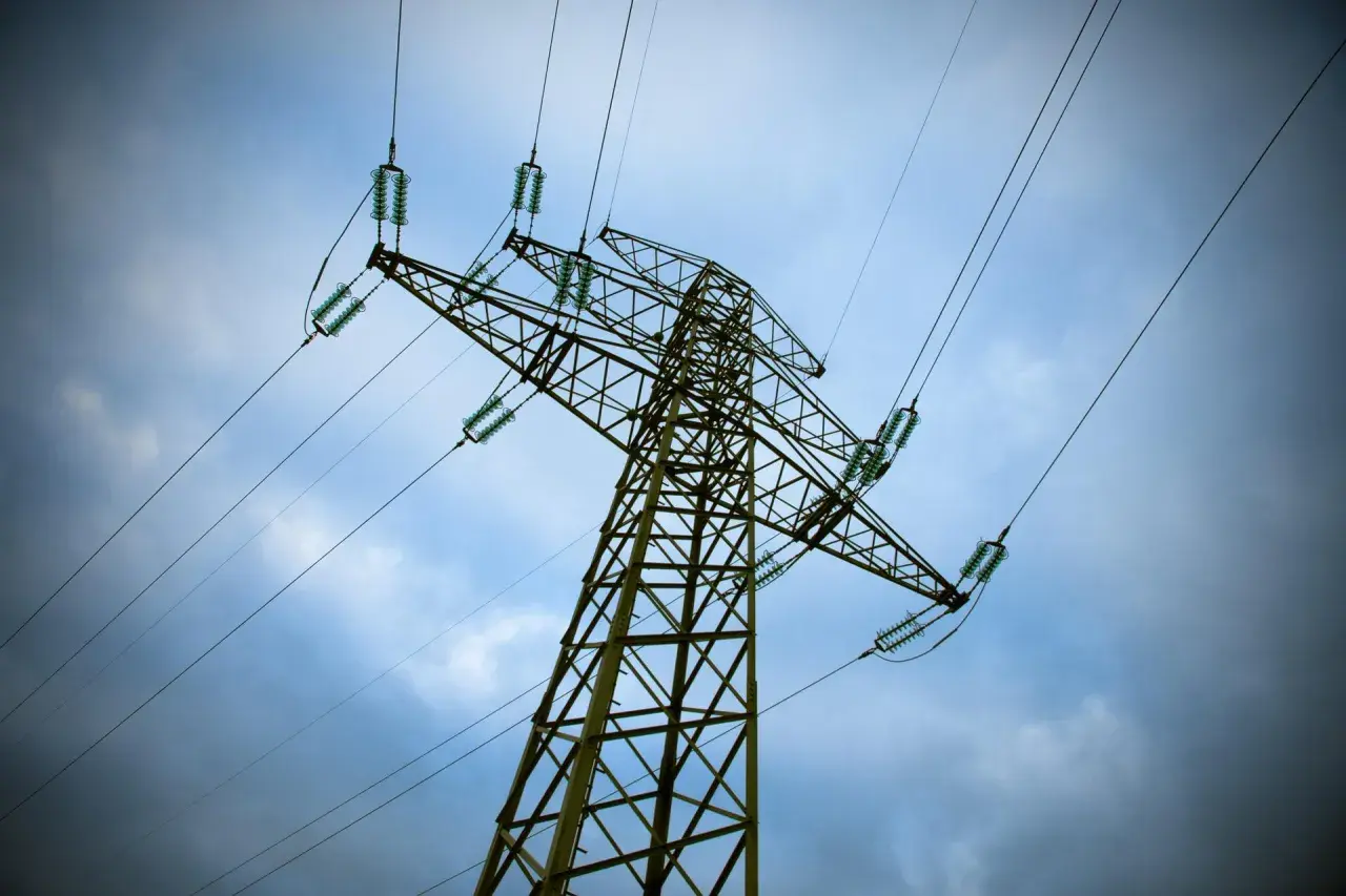 Wysoki słup energetyczny z przewodami, pod niebem pełnym chmur. Mam nadzieję, że nie będzie awarii, bo wtedy czeka nas bonifikata za brak prądu.