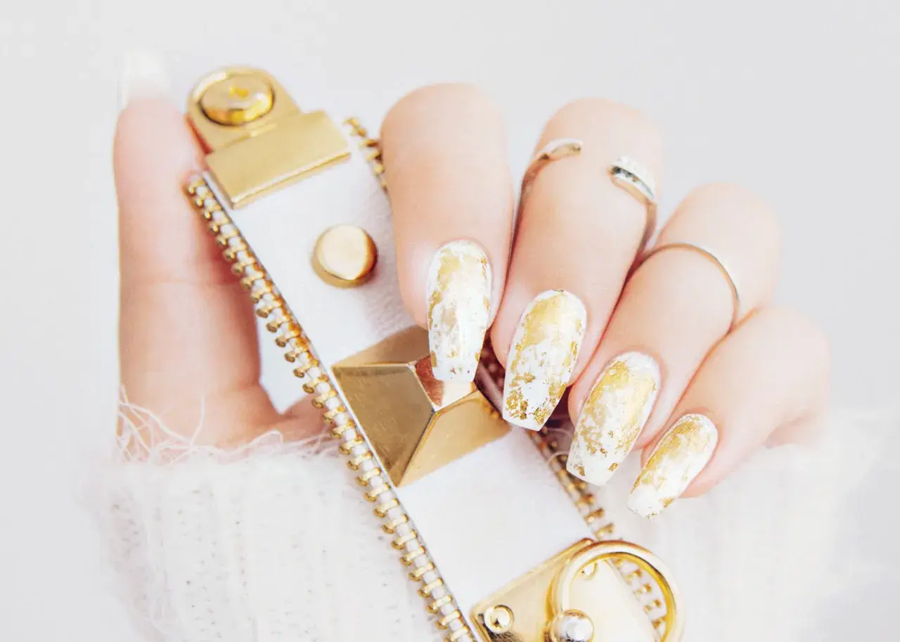 En hand med vita naglar dekorerade med guld nagellack, som en lyxig touch. En vit och guldig accessoar syns också.