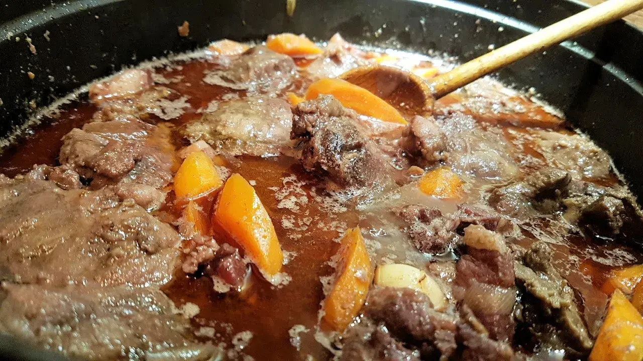 Un plat mijoté de joue de porc avec des carottes, dans une sauce brune savoureuse. Une cuillère en bois remue doucement.
