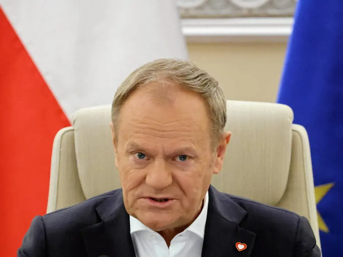 Donald Tusk, z flagą Polski i UE za plecami, w białej koszuli i granatowej marynarce. Czy Donald Tusk ma pochodzenie niemieckie?