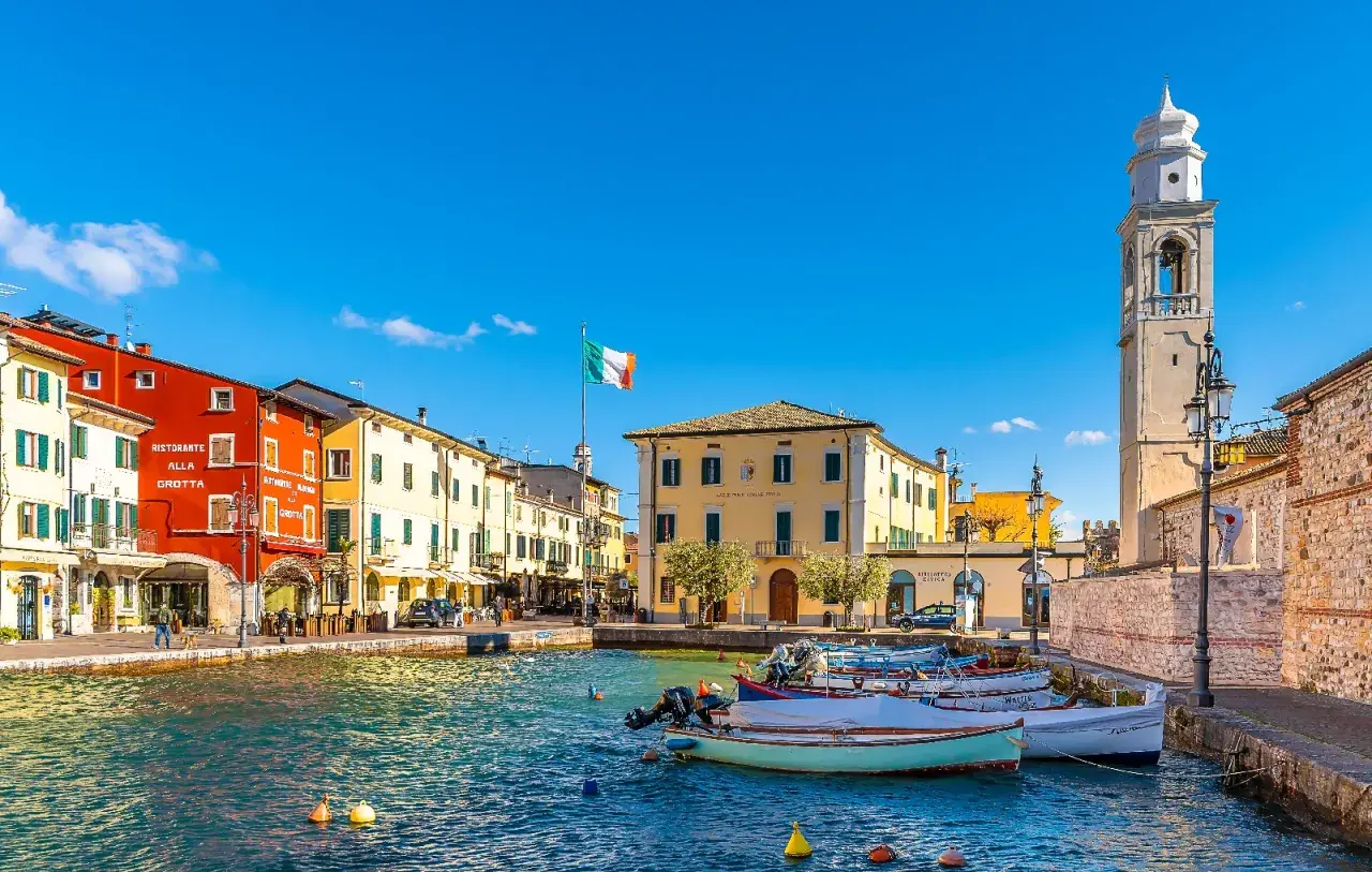 Colorful buildings line a harbor with boats, an Italian flag, and a bell tower under a clear blue sky.