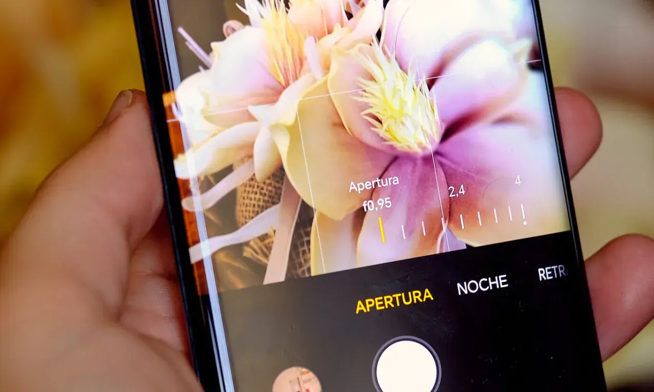Mano sujetando móvil, mostrando interfaz de cámara con flores y ajustes de apertura. Aprende como hacer un selfie con el móvil.