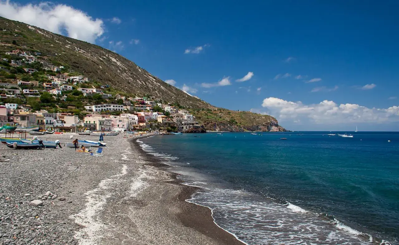 Vue d'une plage de galets sur l'île de Lipari, avec des maisons colorées accrochées à la colline et des bateaux dans la mer d'un bleu profond.