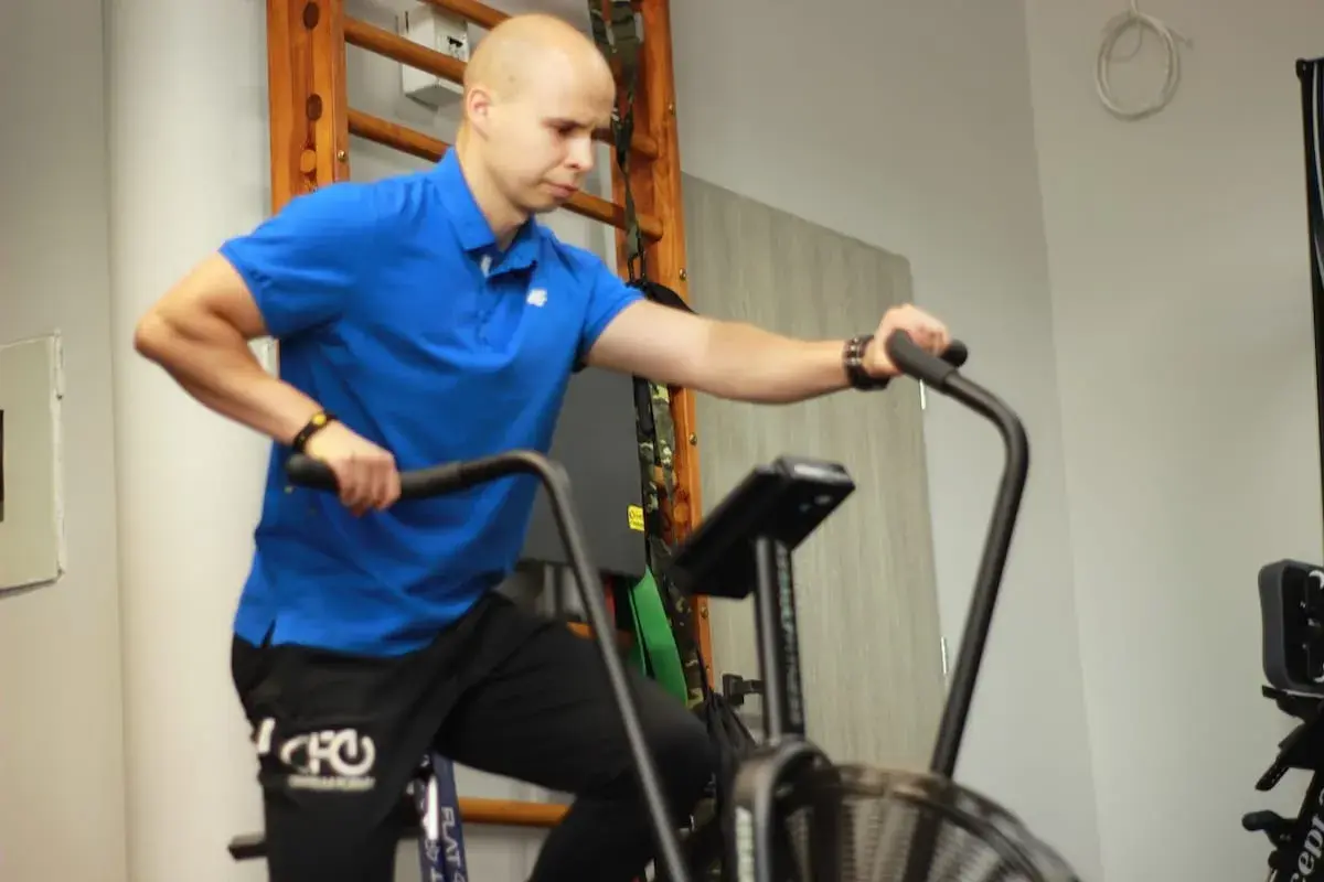 Mężczyzna w niebieskiej koszulce wykonuje ćwiczenia aerobowe na rowerze stacjonarnym, trenując wytrzymałość.