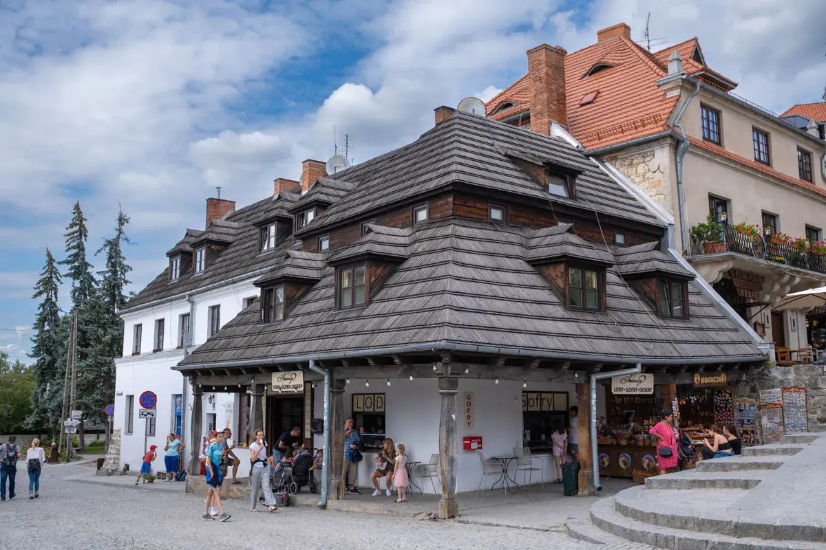 Kazimierz Dolny rynek: urokliwe kamienice z drewnianymi dachami, ludzie spacerują i kupują lody.