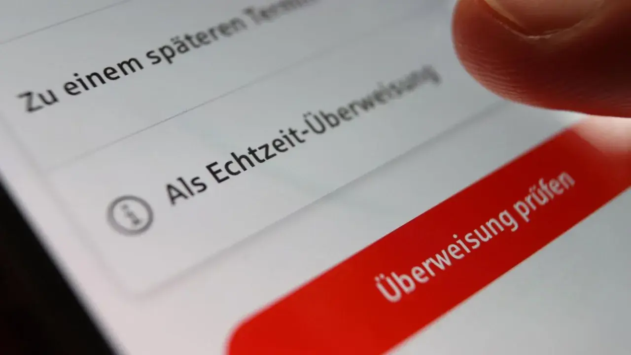 Finger tippt auf "Überweisung prüfen" auf einem Smartphone. Die Option "Als Echtzeit-Überweisung" ist sichtbar. Banken buchen oft sofort.