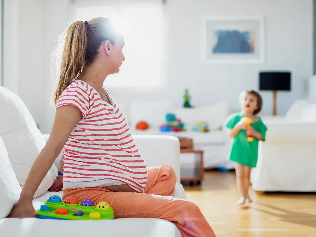 Schwangere Frau sitzt auf Sofa, Kind spielt. Frage: haushaltshilfe nach op wie viele stunden?