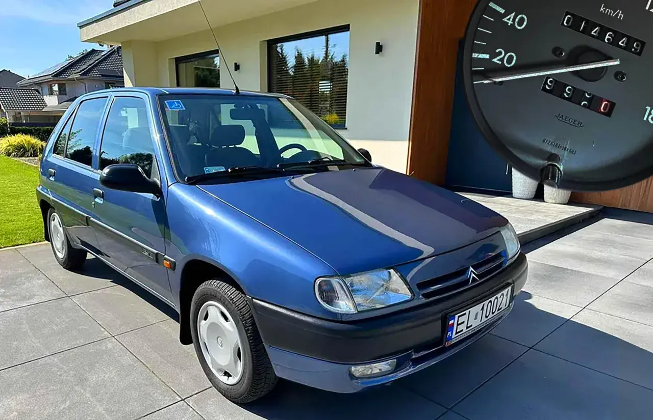 Niebieski Citroen Saxo z niskim przebiegiem, zaparkowany przed nowoczesnym domem. Na desce rozdzielczej widać licznik z przebiegiem 14649 km.