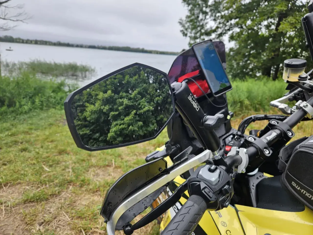 Lusterko motocyklowe z odbiciem drzew i telefonem nawigacyjnym na kierownicy, nad jeziorem.