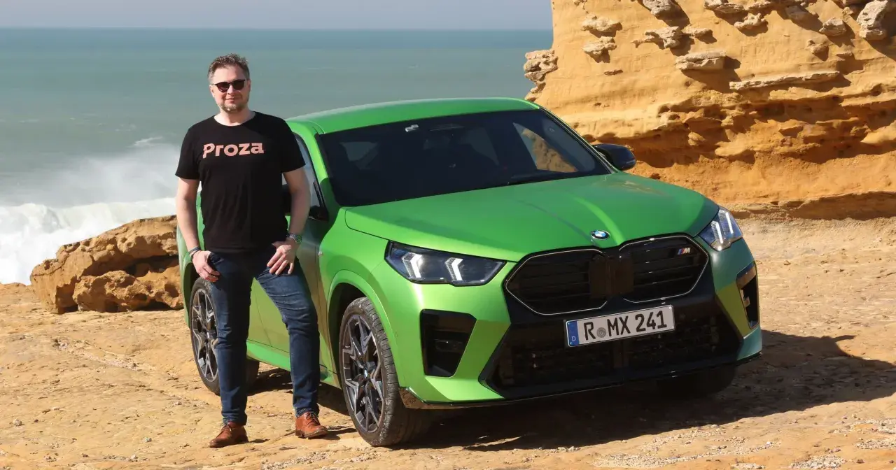 Mężczyzna w czarnej koszulce "Proza" stoi obok jaskrawozielonego BMW X2 M. Samochód zaparkowany jest na skalistym wybrzeżu z widokiem na wzburzone morze.