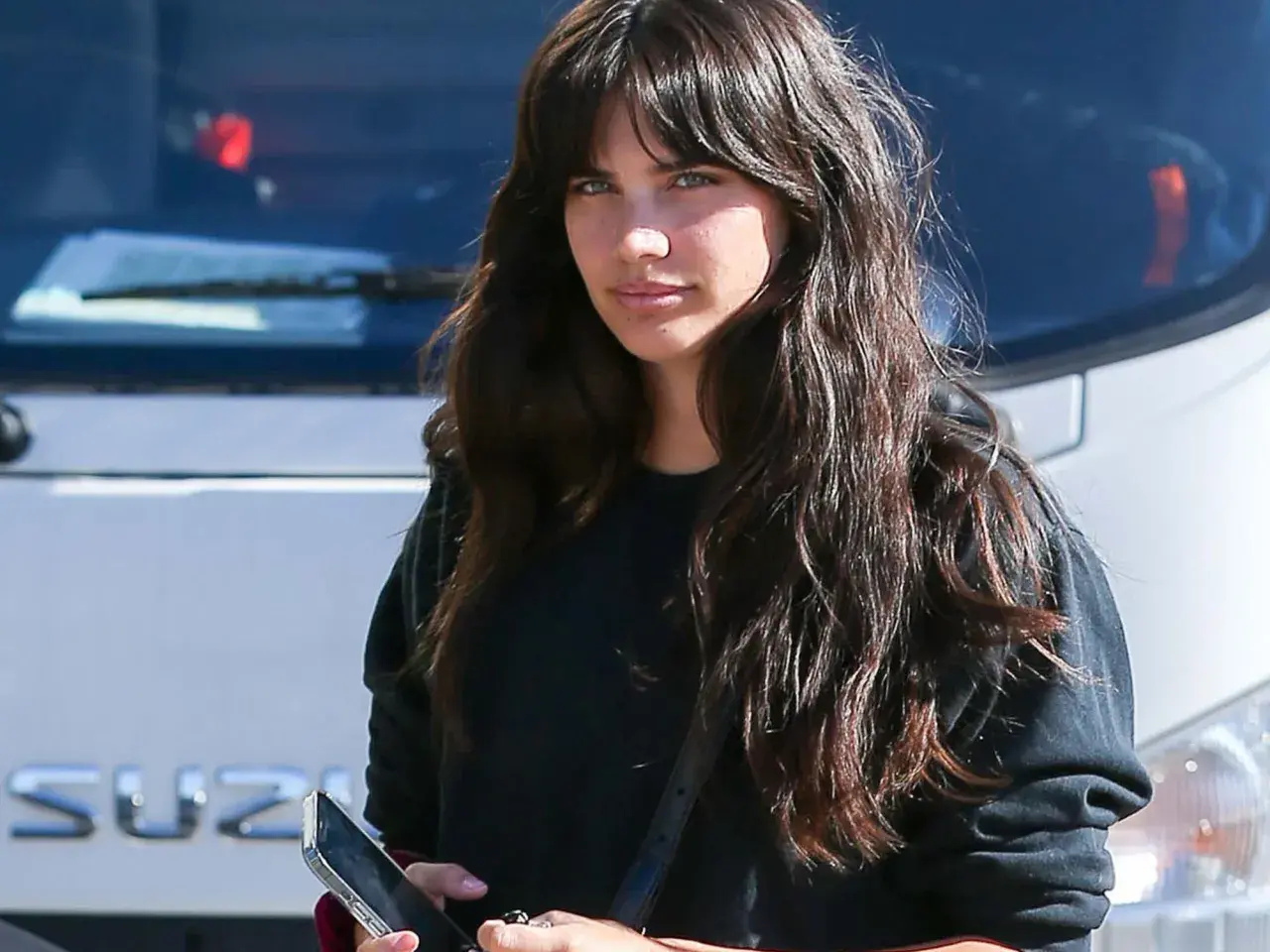 Mujer con cabello largo y oscuro, luciendo un moderno flequillo cortina, mira a la cámara mientras sostiene un teléfono.