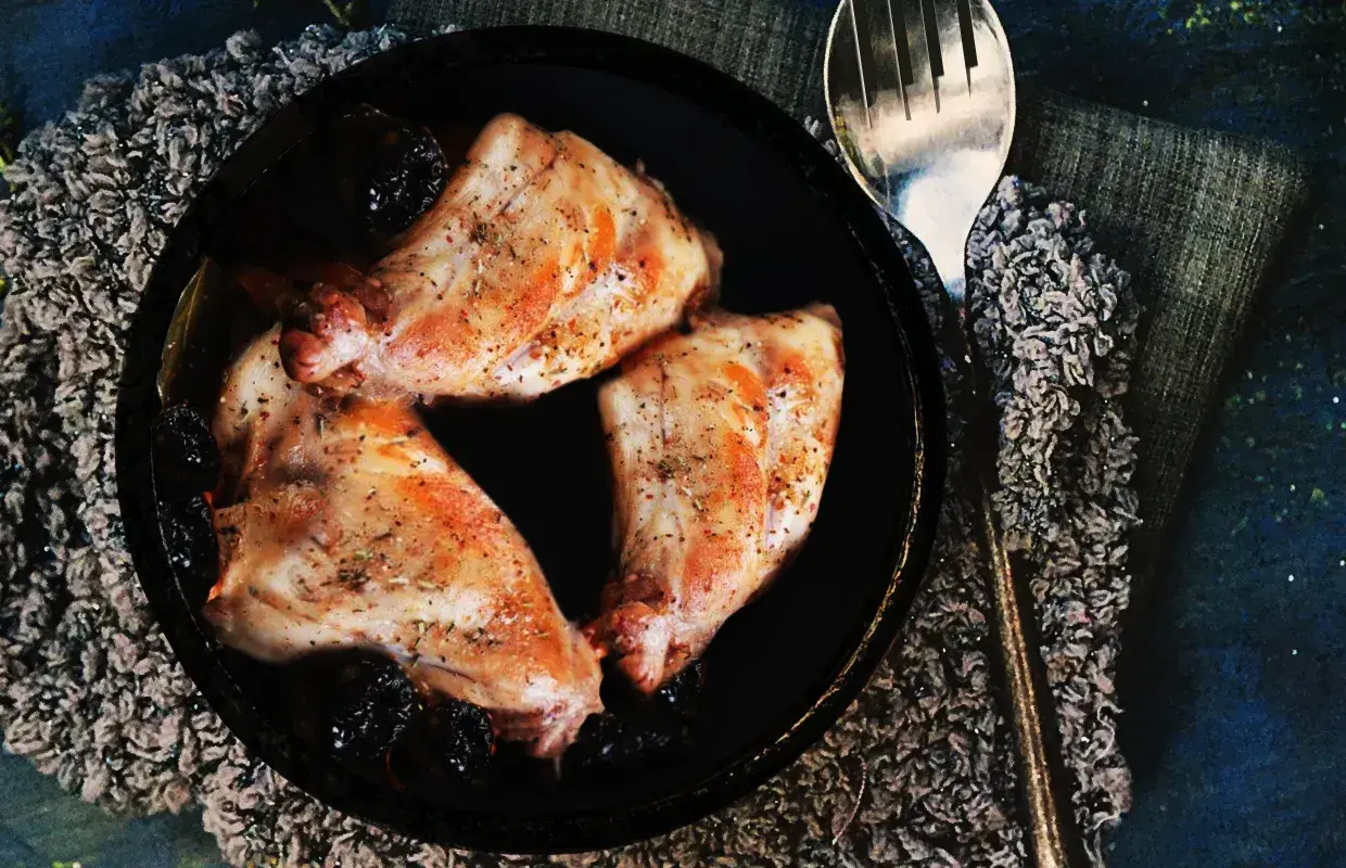 Trois morceaux de lapin aux pruneaux, assaisonnés et dorés, servis dans un plat noir avec une cuillère argentée.