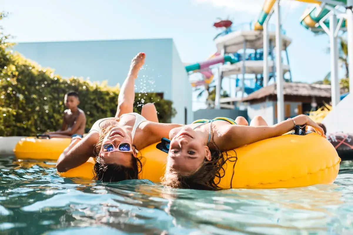 Dwie dziewczyny relaksują się na dmuchanych kołach w basenie. W tle widać zjeżdżalnie wodne, sugerujące, że to największy aquapark w Europie.