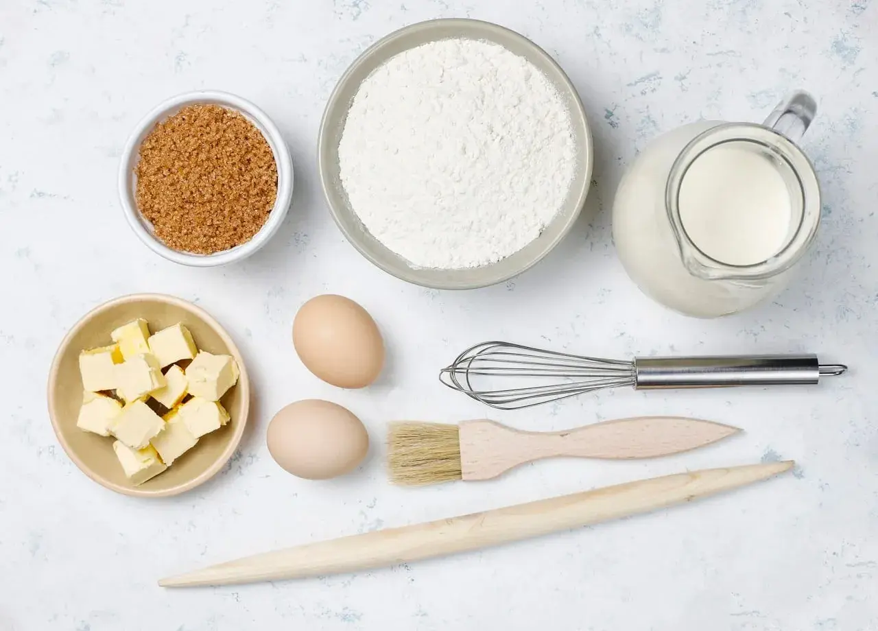 Ingredientes para hornear: harina, azúcar moreno, mantequilla en cubos, huevos, leche en jarra, batidor, brocha y rodillo de madera.